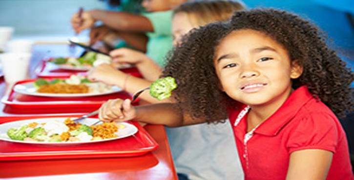 A smiling girl with curly hair sitting at a table, enjoying a meal with broccoli and other dishes, alongside other children.