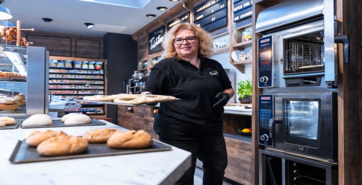 Lächelnde Mitarbeiterin in der Tankstellenbäckerei mit Teigblech vor RATIONAL iCombi Pro Kombidämpfer.