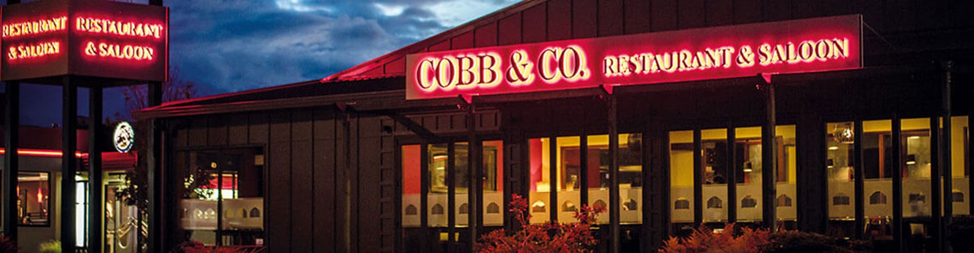Exterior view of Cobb & Co. Restaurant & Saloon at dusk, showcasing bright neon signs and illuminated windows against a darkening sky.