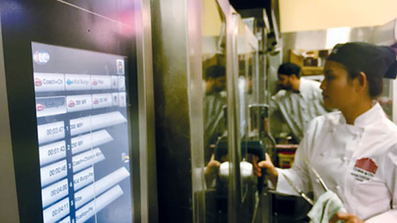 A chef in a white uniform interacts with a Rational oven display system showing cooking times, while other staff work in the background in a commercial kitchen.