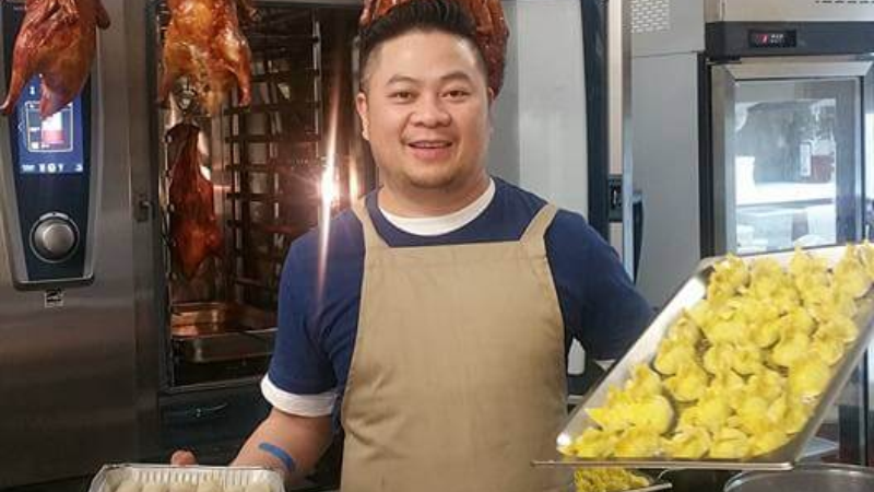 A man in an apron holding a tray of food in a kitchen with hanging ducks and prepared dishes.