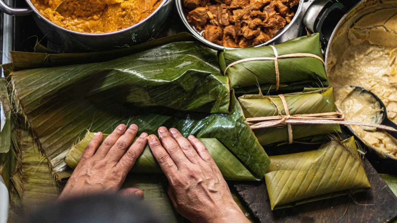 Hands preparing tamales using banana leaves, surrounded by pots of filling and ingredients in a Mexican kitchen.