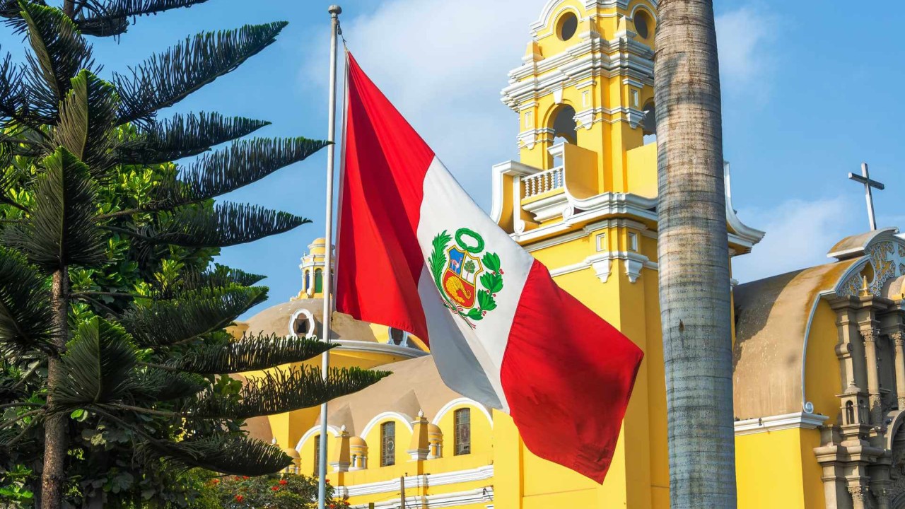 Peruvian flag waving in front of a yellow colonial building with palm trees under a blue sky.