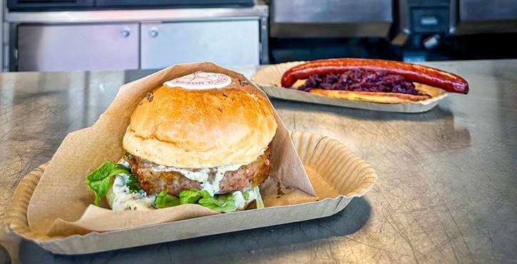 Šťavnatý burger se salátem a omáčkou v papírové misce, v pozadí klobása se zelím.