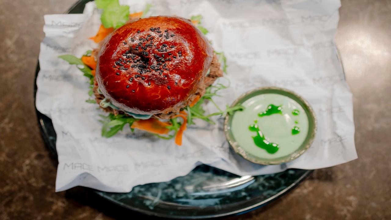 Gurmánský burger s lesklou brioškovou houskou a černým sezamem, podávaný se zelenou omáčkou na černém talíři.