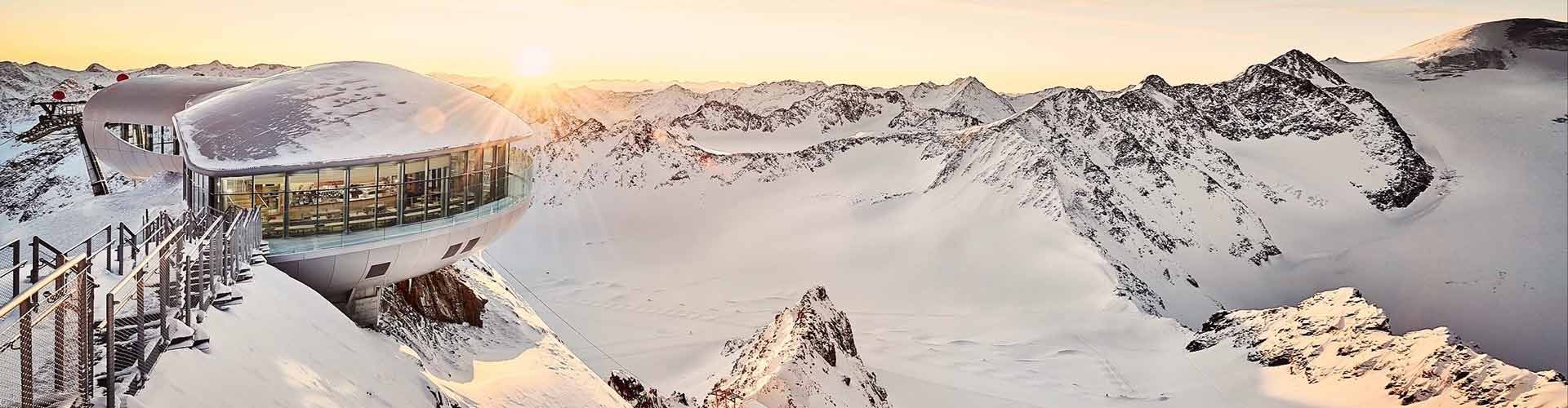 Panoramic view of a snow-covered mountain landscape with a modern observation building and sunset in the background.