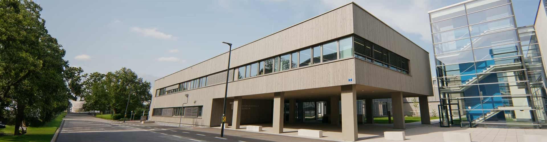 Moderne Architektur der Tourismusschule Klessheim mit verglastem Treppenhaus und Bäumen im Umfeld.