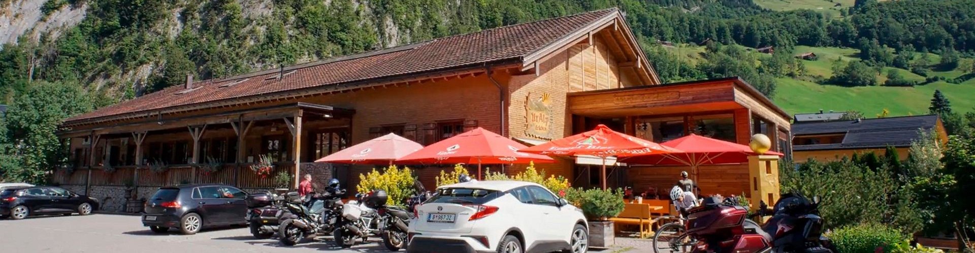 Alpenrestaurant mit roten Sonnenschirmen, geparkten Fahrzeugen und grünen bewaldeten Bergen.