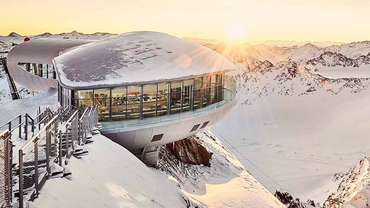 Panoramic view of a snow-covered mountain landscape with a modern observation building and sunset in the background.