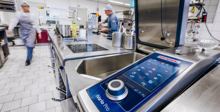 Nahaufnahme des RATIONAL iVario Pro Touchscreens in einer Großküche mit arbeitenden Köchen.