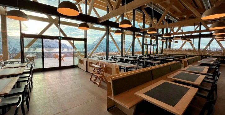 Moderner Innenraum eines Alpenrestaurants mit Holzbalken, Panoramafenstern und Blick auf verschneite Berge.