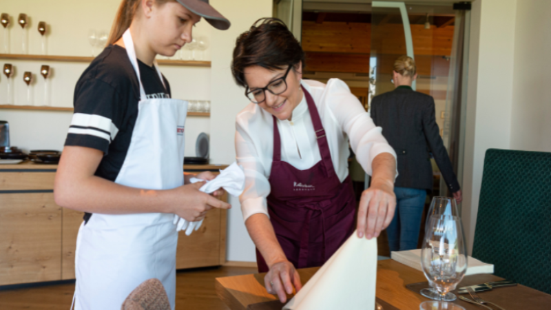 Ausbilderin unterstützt junge Teilnehmerin beim Eindecken eines Tisches im Schulungsraum.