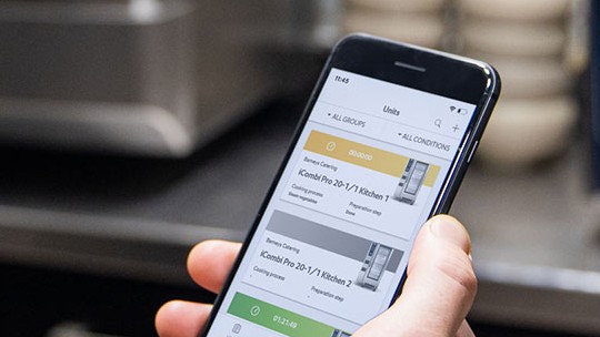 Eine Person hält ein Smartphone auf dem man ConnectedCooking sieht. Im Hintergrund ein iCombi Pro und ein iVario XS.