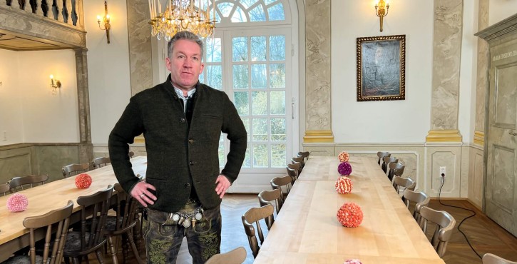 Mann in traditioneller bayerischer Tracht steht in einem eleganten, historischen Speisesaal mit langen Holztischen, die mit bunten Blumenkugeln dekoriert sind.