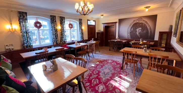 Gemütlicher, traditioneller Gastraum mit Holztischen und -stühlen, bunten Kissen, geblümten Vorhängen und rotem Orientteppich.