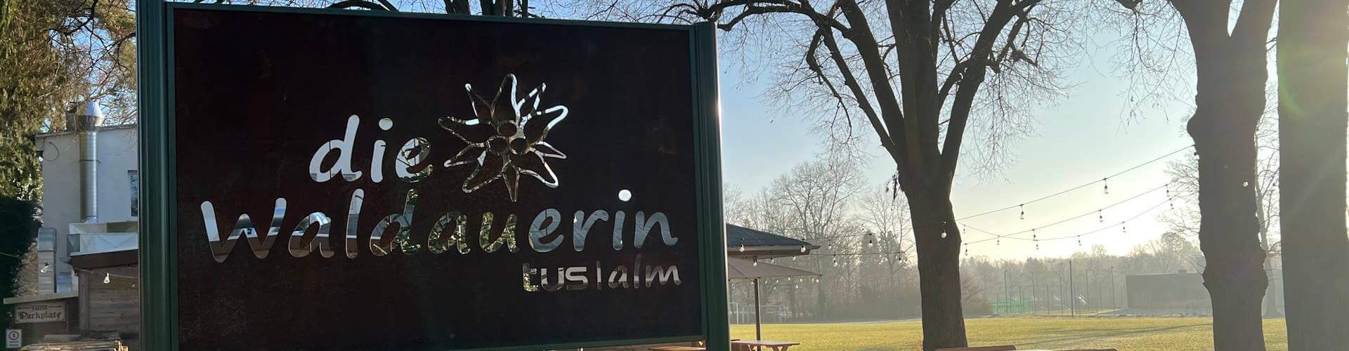 Schild „die Waldauerin – TUSI Alm“ im sonnigen Biergarten mit Holzbänken, Bäumen und Lichterketten.