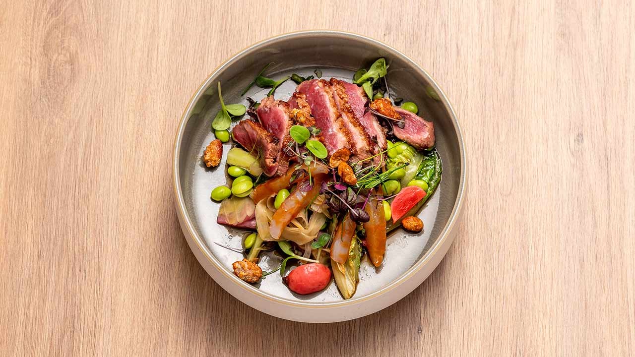 Modern angerichtetes Restaurantgericht mit aufgeschnittenem, angebratenem Fleisch, frischem Gemüse und Microgreens auf Holztisch.