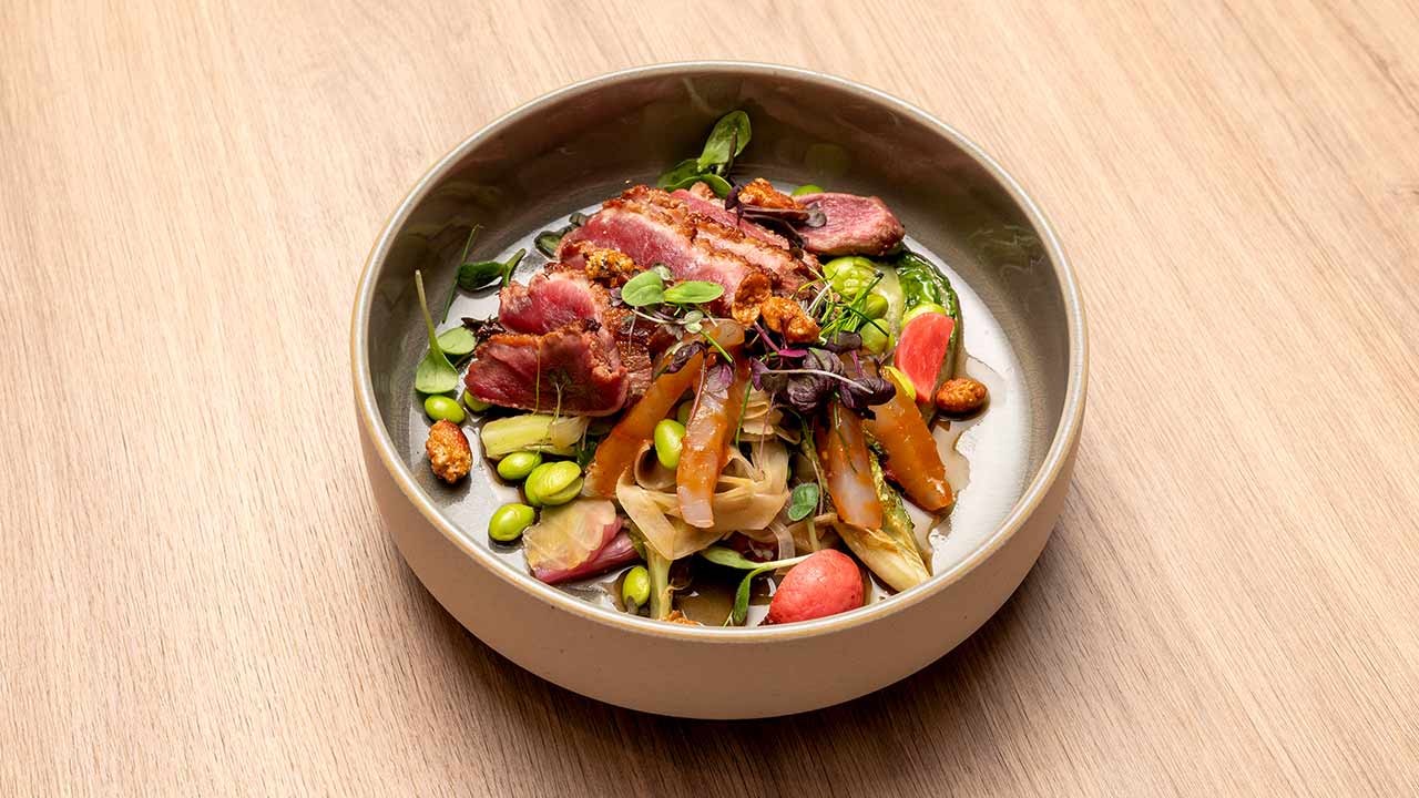 Moderne Restaurantschale mit aufgeschnittenem, angebratenem Fleisch, Edamame, saisonalem Gemüse und Microgreens auf Holztisch.