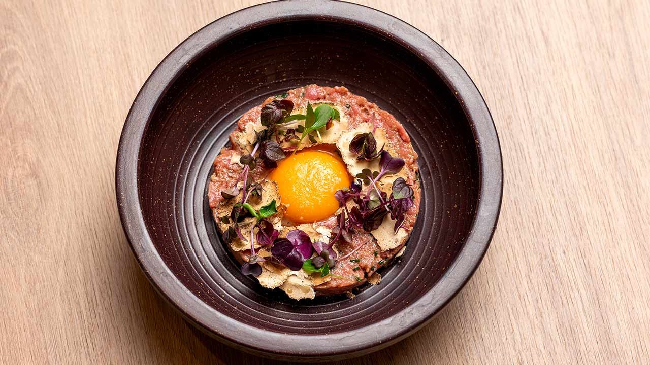 Rindertatar mit rohem Eigelb, gehobeltem Käse und Microgreens in dunkler Schale auf Holztisch.