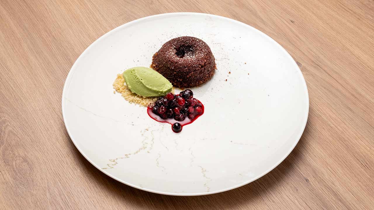 Schokoladenküchlein mit grünem Eis, Beerenkompott und Streuseln auf weißem Teller.
