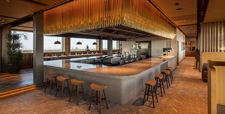 Moderne Showküche im Restaurant mit großem Tresen, Barhockern und goldener Deckenbeleuchtung.