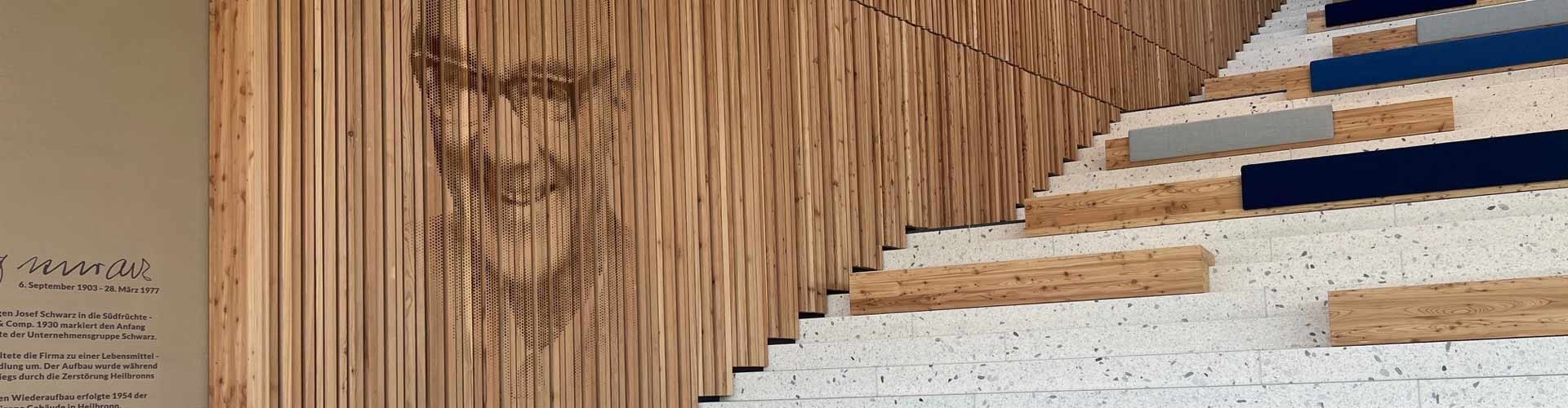 Innenraum mit Holzlamellenwand und eingraviertem Porträt sowie gestufter Sitztreppe in modernem Schulgebäude.