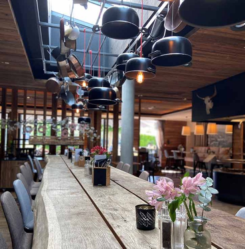 Rustikaler Holztisch mit Blumenvasen unter hängenden Töpfen und warmem Licht im Restaurant.