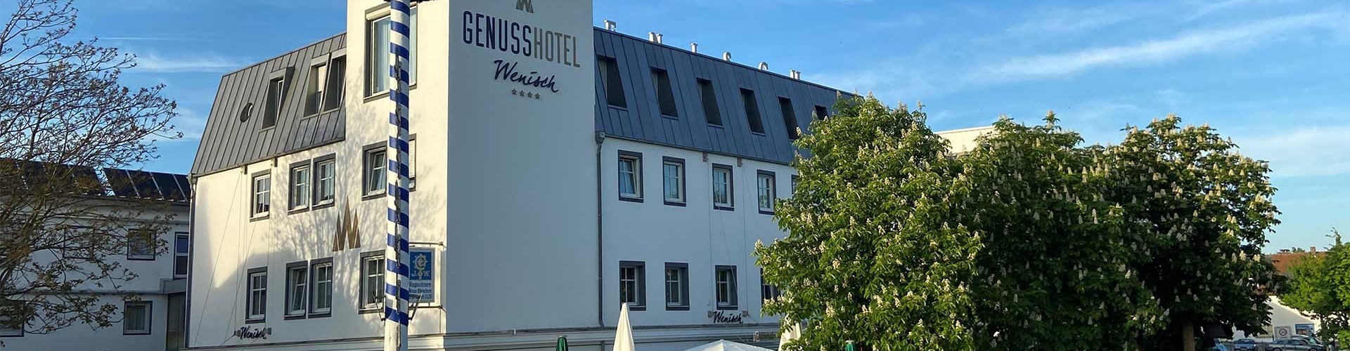 Außenansicht des Genusshotel Wenisch mit blau-weißem Maibaum und Bäumen.