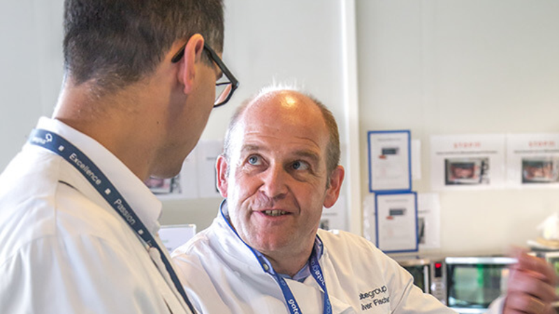 Two colleagues in chef jackets talking in a professional kitchen setting.