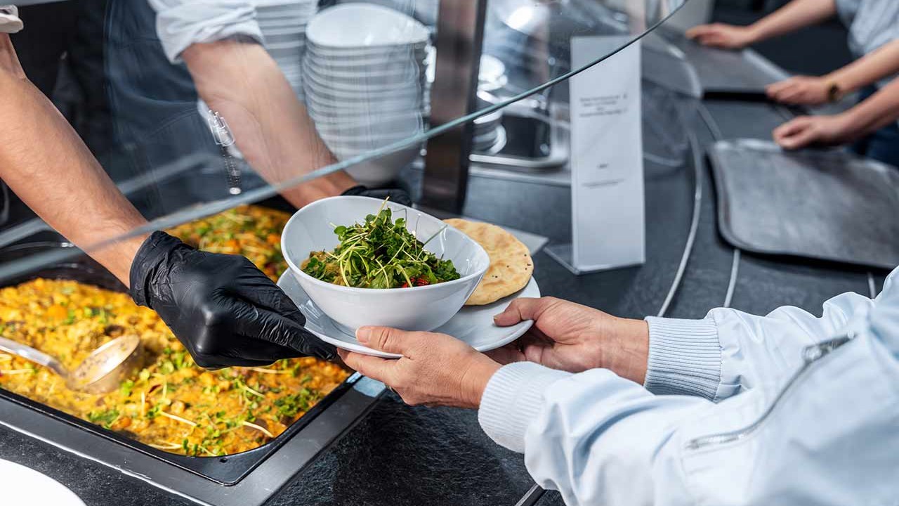 Mitarbeiter reicht frische Salatschüssel an Gast am Ausgabebereich in Profiküche.