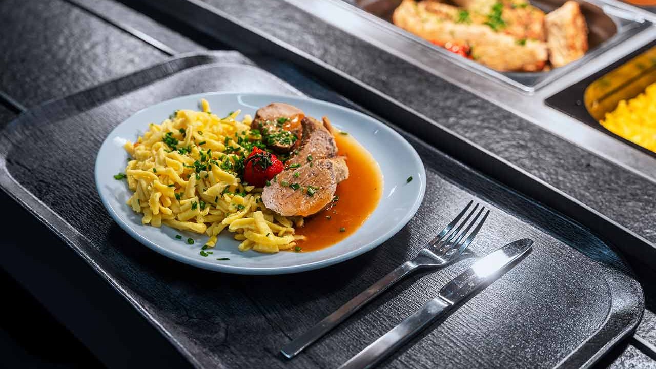 Teller mit Fleisch in Sauce, Spätzle und Kräutern auf Buffettheke.