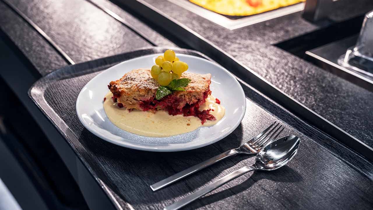 Stück Beeren-Streuselkuchen mit Vanillesauce und Trauben auf Buffettheke.