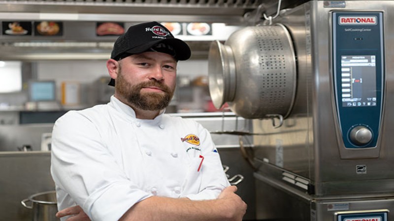 A chef in a white jacket and a black hat branded with "Hard Rock Cafe" stands confidently in a commercial kitchen with arms crossed, next to a RATIONAL SelfCookingCenter appliance.