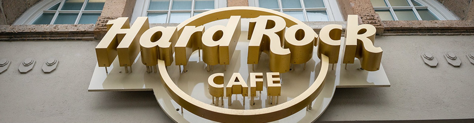 Exterior view of a Hard Rock Cafe building, showcasing the bold, golden 3D logo mounted on a textured stone facade with a large arched window above the sign.