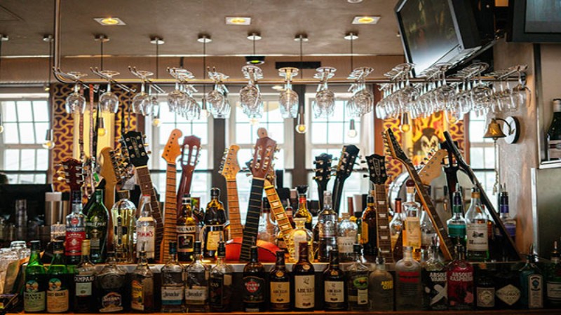 A lively bar scene featuring an array of liquor bottles on the counter, with multiple guitar necks displayed overhead in place of taps, and glassware hanging from the rack above, set against a backdrop of intricate, colorful patterns.