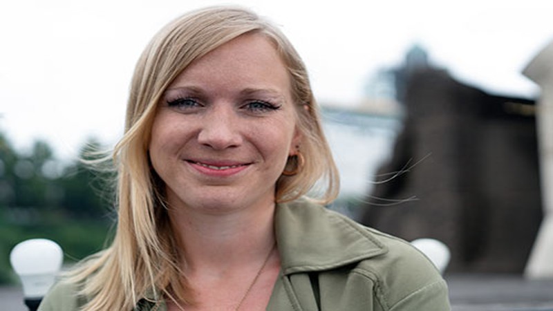 Close-up of a smiling individual with blonde hair, wearing a green jacket, with a blurred urban background.