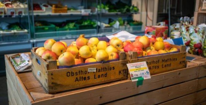 Holzkisten mit Äpfeln im HOBENKÖÖK Markt vor Regalen mit frischem Gemüse.