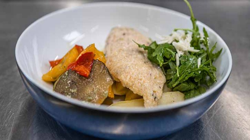 Schale mit Fisch, gegrilltem Gemüse und Rucolasalat auf einer Küchentheke.