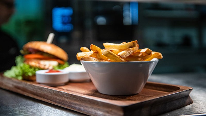 Schale mit Pommes auf einem Holztablett mit Burger im Hintergrund.