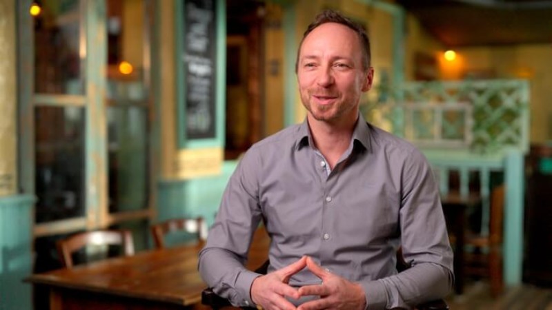 A man in a gray shirt smiling and sitting with his hands clasped in a restaurant setting.