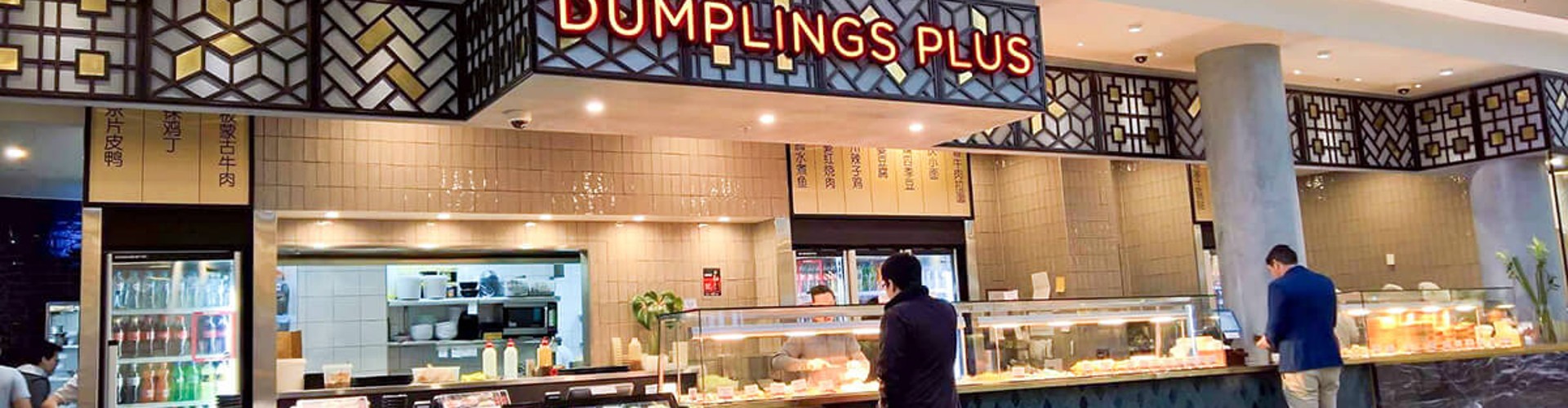 Interior of a food court featuring "Dumplings Plus" restaurant with customers browsing and two individuals standing at the counter.