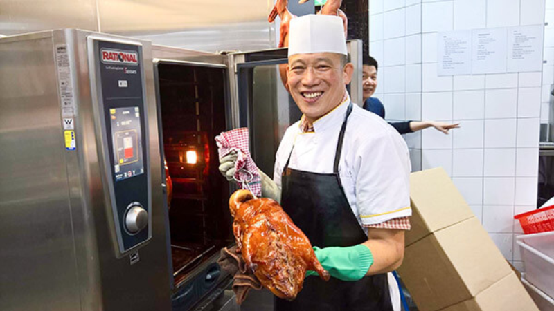 A smiling chef wearing a white hat and apron holds a roasted chicken in front of a Rational oven, with another person visible in the background.