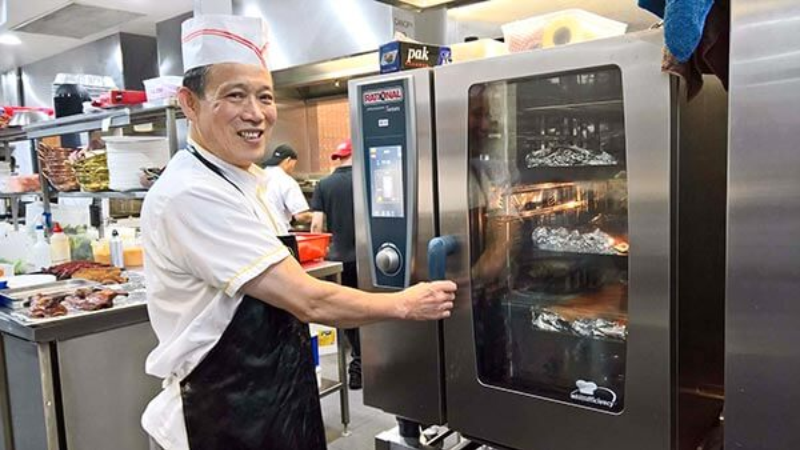 A chef wearing a white uniform and headband smiling while opening an Rational oven in a busy kitchen.