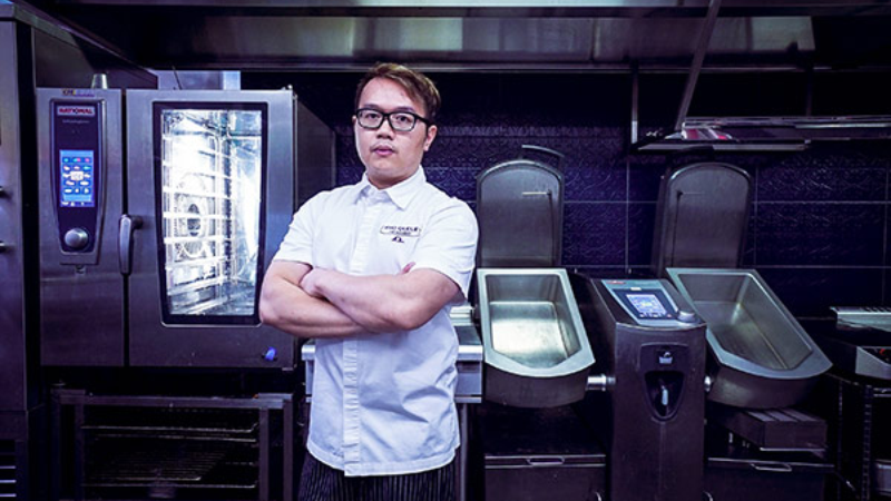 A chef stands with arms crossed in a commercial kitchen, wearing a white chef‘s coat with the logo "Culinary Institute of America." The background features stainless steel Rational kitchen equipment.