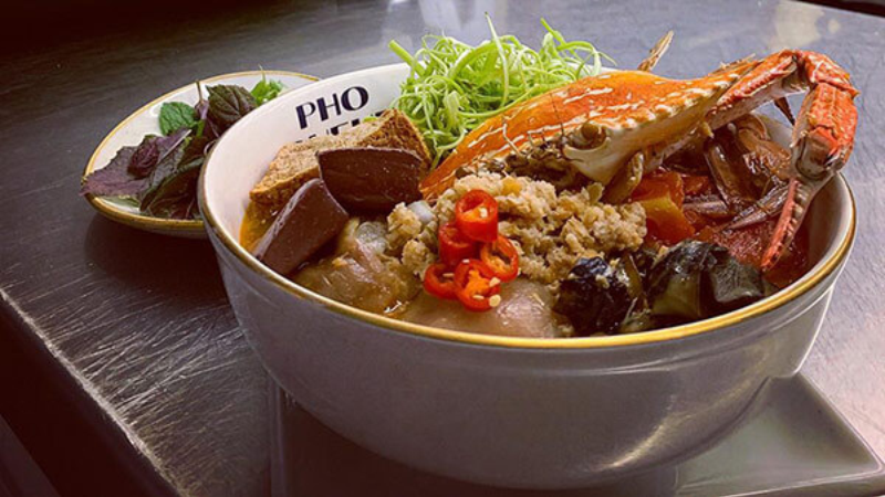 A bowl of pho with crab, garnished with chili peppers, herbs, and lime, served alongside fresh greens.