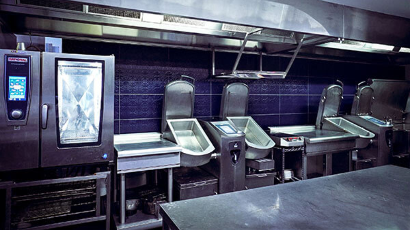 Interior of a commercial kitchen featuring stainless steel appliances, including 2 iVario Pro, a Rational oven, and a streamlined workspace under blue lighting.