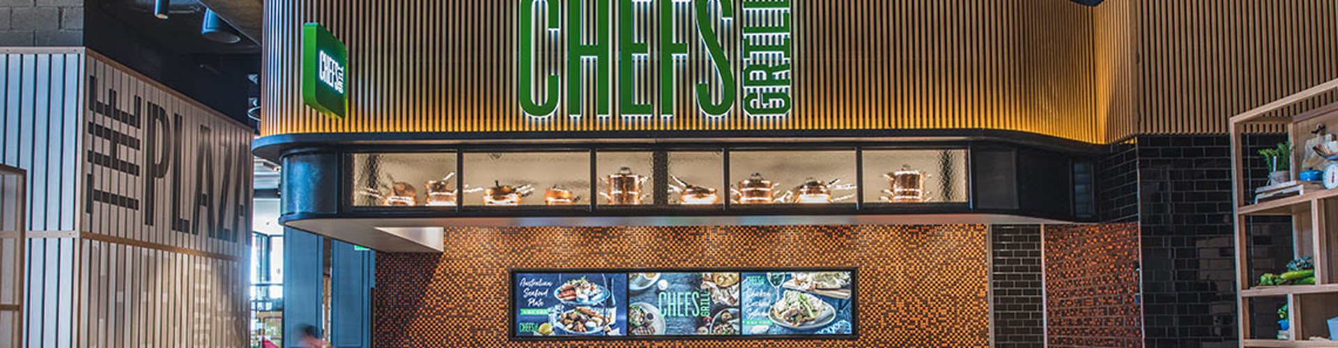 Interior of a modern CHEFS restaurant with a prominent green logo above the service counter, digital menu displays, and stylish seating area. The decor features wood shelving with various items, a patterned tile floor, and industrial-style lighting.