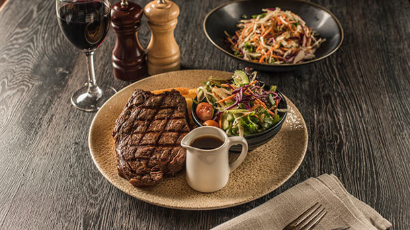 A plate of grilled steak served with a side of mixed vegetables and a small jug of sauce, accompanied by a salad and a glass of red wine on a wooden table.