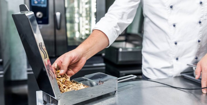 Chef in white jacket adding wood chips to a smoker device in a commercial kitchen, with a RATIONAL iCombi Pro oven behind.