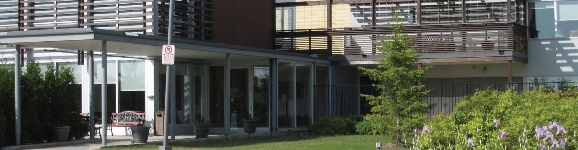 A modern building with a curved driveway, a garden with flowers, and large windows with a shaded balcony.